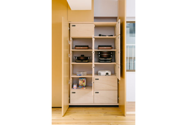Tall wooden kitchen cabinet with doors open, showing pans and skillets on multiple shelves, a small white projector, and children's books on the bottom shelf/ drawers.