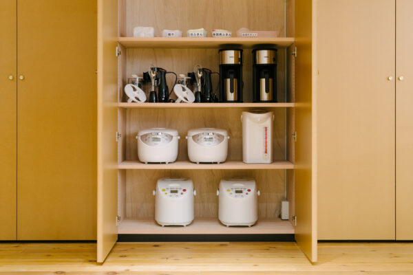 Open wooden cabinet with four shelves holding small kitchen appliances: labeled containers on the top, two black coffee makers and a white kettle on the second shelf, and multiple rice cookers on the lower shelves.