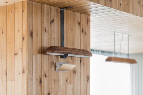 2017_0303_0049 Wood-paneled wall with a curved wooden wall light fixture and a white rectangular sconce beneath it in a sunny interior.