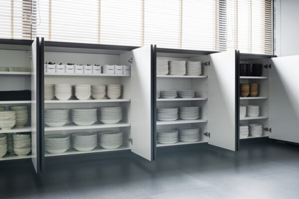 2017_0303_0056 Open kitchen cabinets neatly filled with stacked white plates, bowls, and cups on a gray floor under window light.