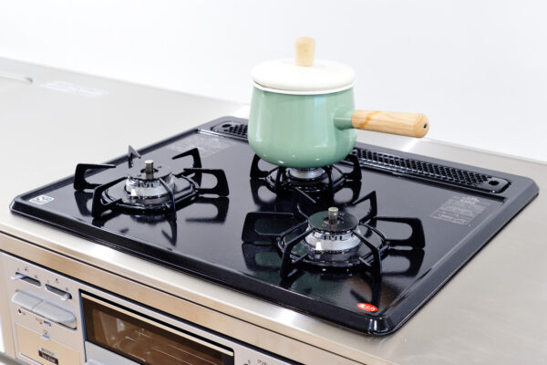Green enameled pot with a wooden lid knob sits on a gas stove burner on a stainless steel countertop stall.