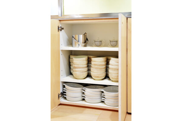 Open kitchen cabinet with three shelves: stacked beige bowls on the middle and bottom shelves; top shelf has a metal bucket and glass teacups.