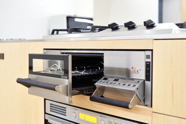 Built-in stainless steel oven with its door open, showing metal racks inside, mounted in light wood cabinetry.
