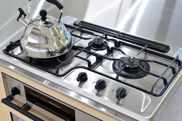 Stainless steel gas stove with a shiny kettle on the left burner and a built-in oven beneath, reflecting the kitchen surroundings.