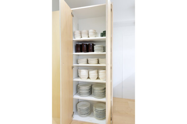 Open tall kitchen cabinet with shelves of white bowls and plates; dark brown mugs on one shelf and small green plates on the top shelf.