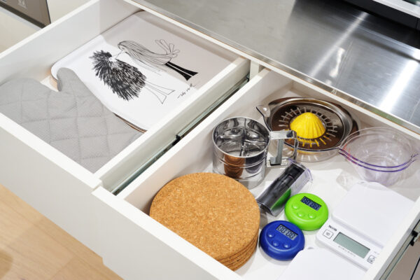 Open kitchen drawer showing baking and kitchen tools: cork coasters, metal measuring cups, juicer, scale timers, and a white digital scale cabinet organizer.