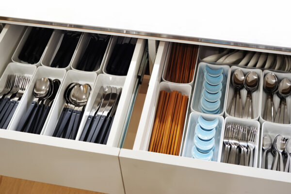 Drawer of organized cutlery: forks, knives, and spoons in separate white compartments, with blue-handled spoons and orange chopsticks visible at the center.