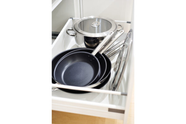 Stack of black nonstick frying pans and lids in a kitchen drawer, with stainless-steel handles visible