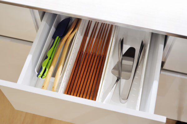 Open kitchen drawer showing utensils in a white organizer: green silicone-tipped tongs and a blue spatula on the left, wooden spoons in the middle, and stainless steel forks and a spatula on the right.