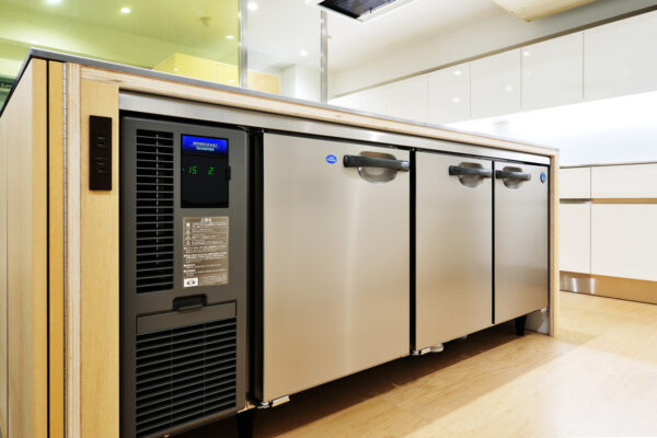Under-counter stainless steel refrigerators with black control panel and handles, set in a light-wood cabinet in a modern kitchen.