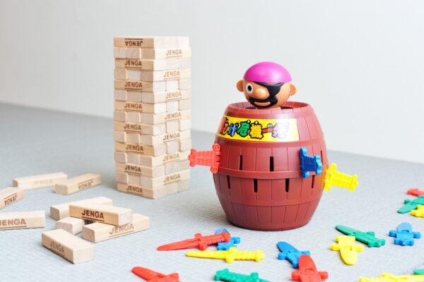 A Jenga tower with scattered blocks sits next to a red pirate-themed barrel toy with a pink hat popping out the top, surrounded by colorful plastic swords and pieces.