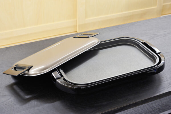 Open metal lunch box with hinged lid lying on a dark wooden table, light wood-paneled wall in the background.