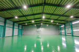 Spacious empty warehouse with a glossy green floor, teal support columns, and a green metal roof lit by bright overhead lights, seen from the center toward a distant doorway.