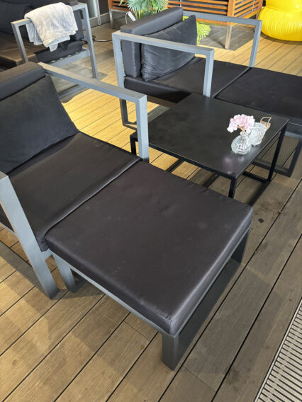 IMG_1067 Outdoor lounge with black cushioned chairs and a small black table holding a glass vase of pink flowers; wooden deck.
