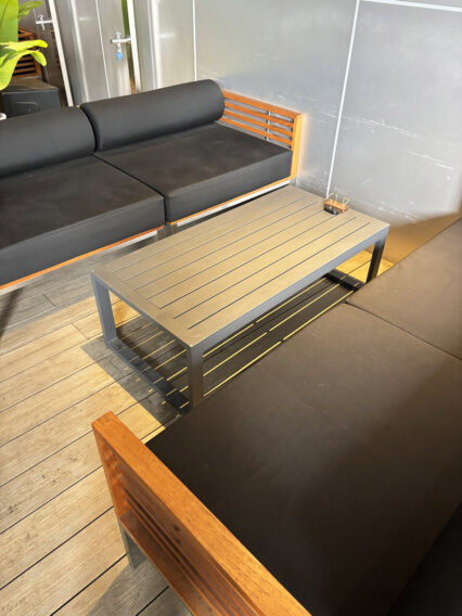 IMG_1068 Outdoor seating area with black cushioned sofa and matching cushions, wooden slatted backrest, and a metal-framed rectangular coffee table on a wooden deck.