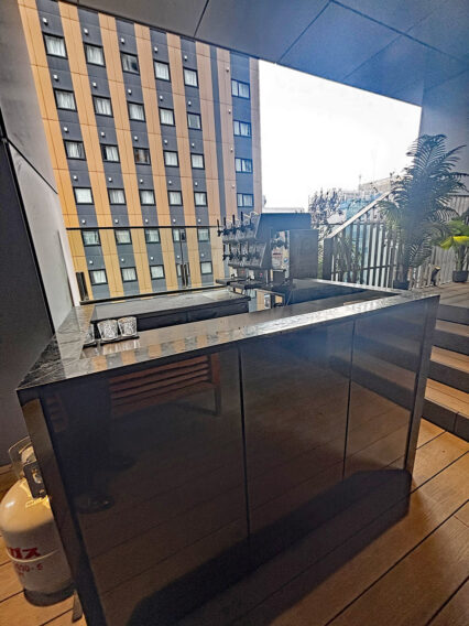 IMG_1069 Rooftop bar area with a dark marble counter and drink dispenser, overlooking a modern orange-gray apartment building.