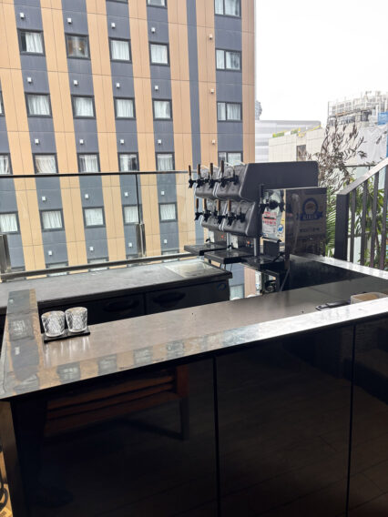 IMG_1070 Outdoor bar counter with a multi-tap beer dispenser and two crystal glasses, overlooking a city building with orange and gray panels
