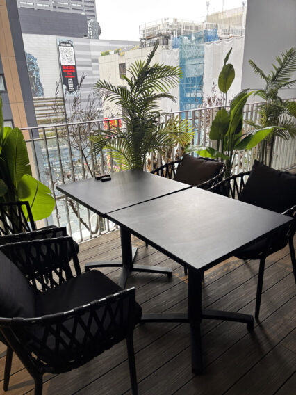 IMG_1072 Outdoor balcony seating area with a black square table and woven chairs, surrounded by tropical plants and a city skyline in the background.