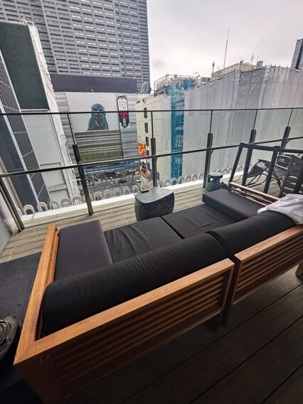 IMG_1073 Rooftop balcony with black cushioned seating and a wooden frame, overlooking a city street with tall buildings.