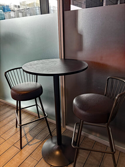 IMG_1075 Round black pedestal table with two metal-bar stools on a wooden floor beside frosted glass panels at a cafe corner.
