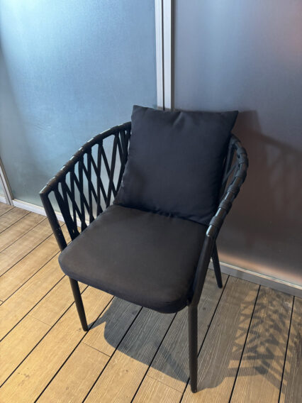 IMG_1076 Black woven patio chair with a dark cushion on a wooden deck by frosted glass panels.