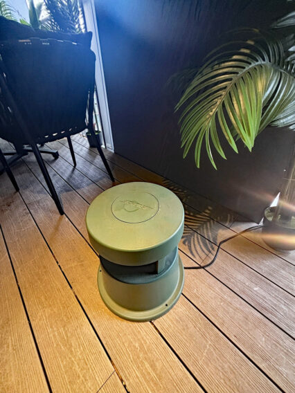 IMG_1083 Round green circular stool on a wooden deck beside a black chair, with a potted plant in the background and a power cord nearby.