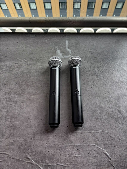 IMG_1085 Two black Shure handheld microphones lying side by side on a gray stage floor with a railing and building visible in the background.