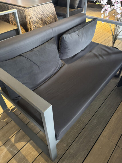 IMG_1088 Outdoor dark gray metal-framed sofa with black cushions on a wooden deck, next to a flowering plant arrangement.