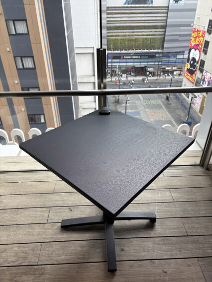 IMG_1089 Square, diamond-oriented outdoor table with a textured dark top on a balcony, overlooking a city street with tall buildings and a cinema sign below.