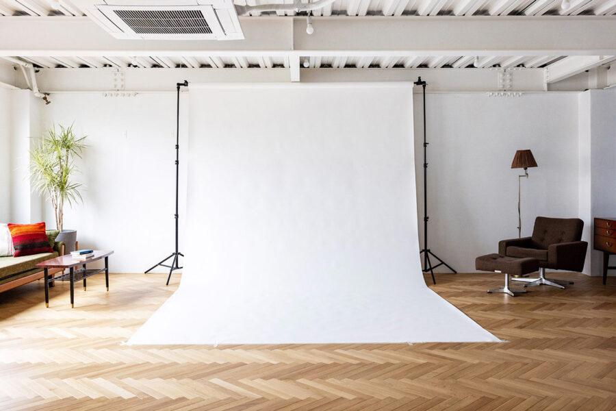 Bright photography studio with a white seamless backdrop, a sofa and coffee table on the left, and a brown lounge chair with a lamp on the right.