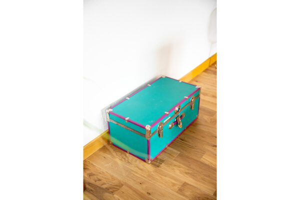 Teal vintage luggage trunk with pink trim and metal clasps on a wooden floor near a white wall.
