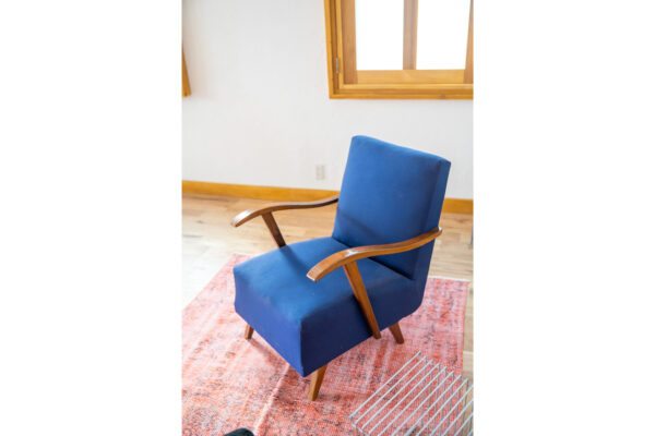 Blue upholstered armchair with curved wooden arms sits on a pink rug in a bright room by a window.