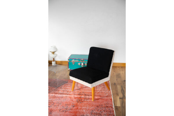 Woven chair with a black cushion sits on an orange rug, with a turquoise case and metal wire table in a bright room.