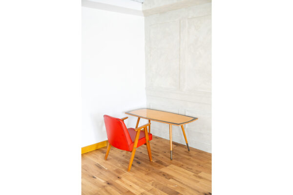 Red upholstered chair with wooden legs beside a light wooden desk in a bright minimalist room.