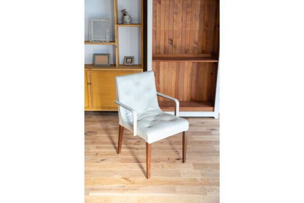 White upholstered armchair with wooden legs, placed on a wooden floor beside a tall shelving unit and cabinet in a sunlit room.