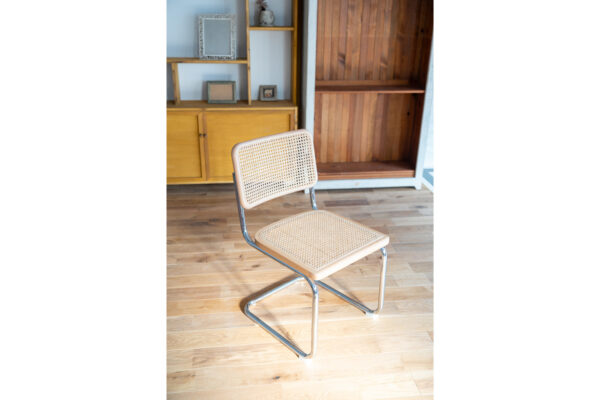 Beige caned chair with metal legs on a wooden floor, in a room with wooden shelves and cabinet behind it.