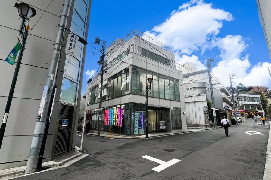 IMG_45940 Modern urban street with a glass-fronted building, neon signs, and a bright blue sky. Pedestrians walk along the paved road with street markings visible.",