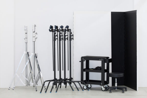 Studio with tall light and grip stands, a rolling cart with cables, and a black adjustable stool in a white room.