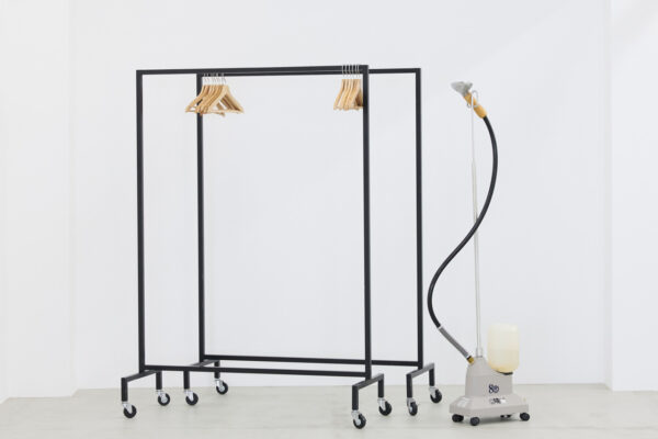 Black metal rolling clothes rack with several wooden hangers, positioned next to a tall garment steamer system on the right side of a white room.