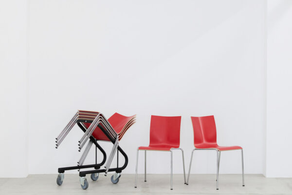 Stacked red chairs on wheeled bases beside two red chairs in a bright white room, showing a simple seating area.
