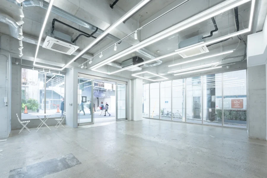 Industrial lobby with exposed ceiling, linear LED lights, and glass walls; small white table and chairs by the window near the entrance.